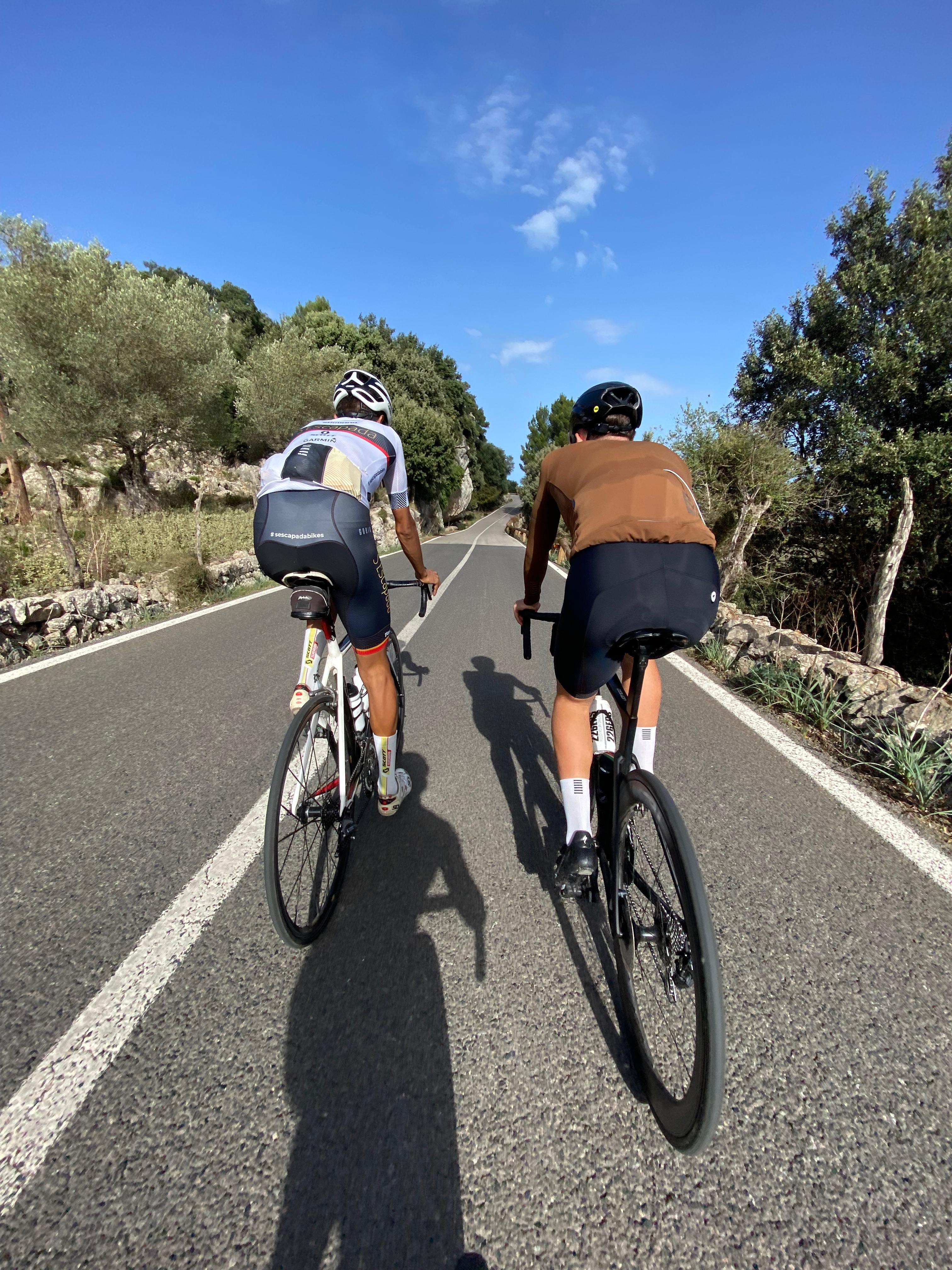Thomas and Xavier riding side by side on a scenic Mallorcan road lined with olive trees and stone walls under a clear blue sky