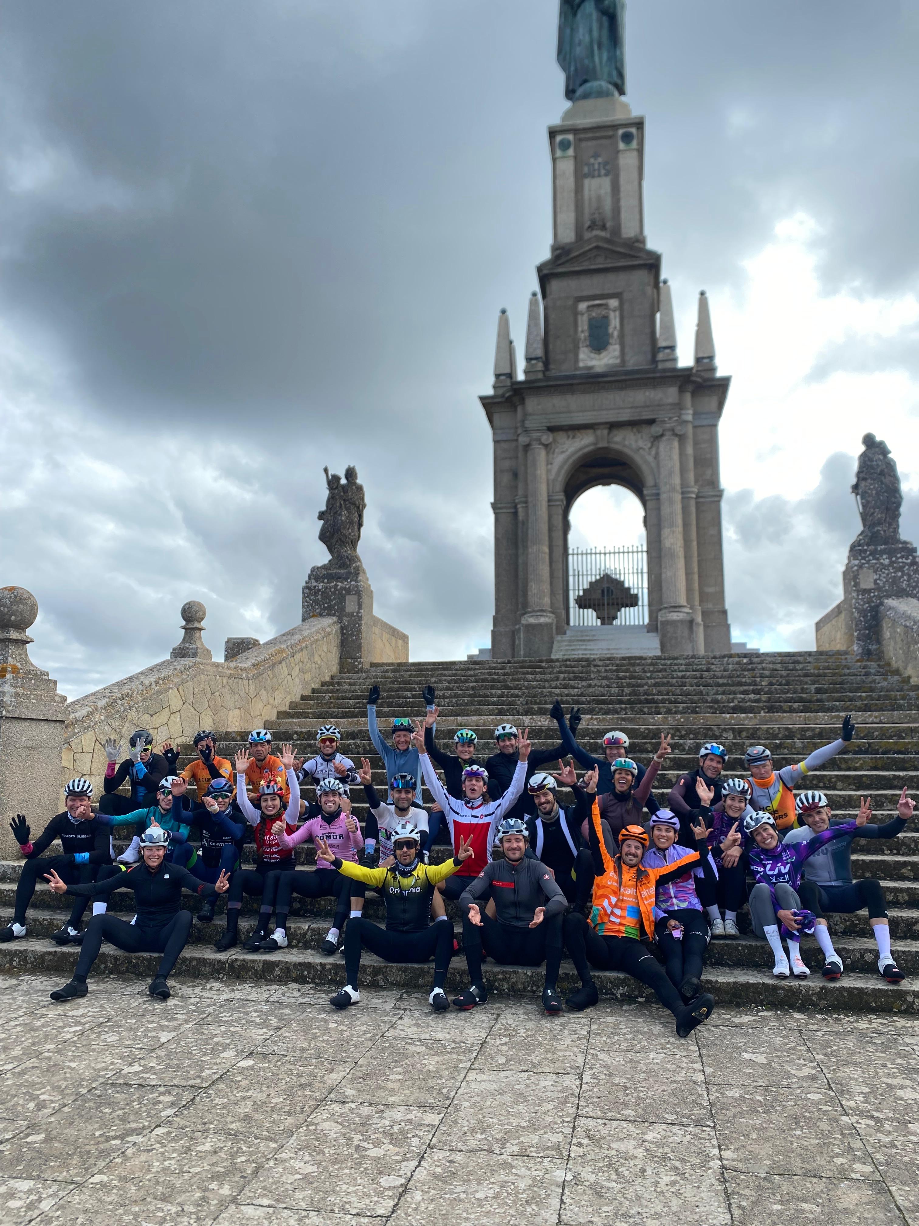 Large group of cyclists celebrating at the top of Puig de Sant Salvador, Mallorca, posing on the stone steps of the hilltop sanctuary with arms raised