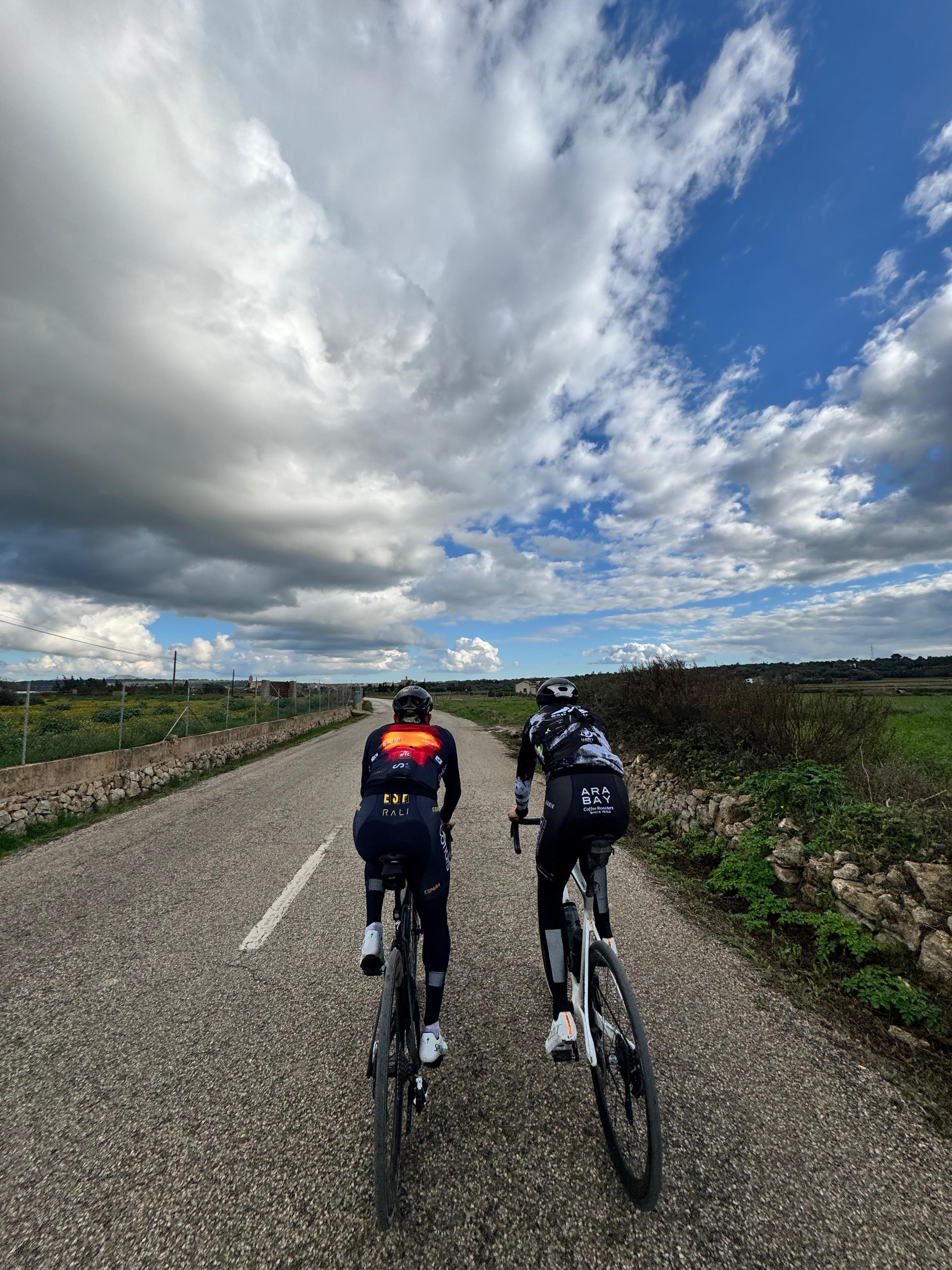Xavier Cañellas and a client riding side by side on a quiet Mallorcan country road, under a dramatic blue sky with scattered clouds