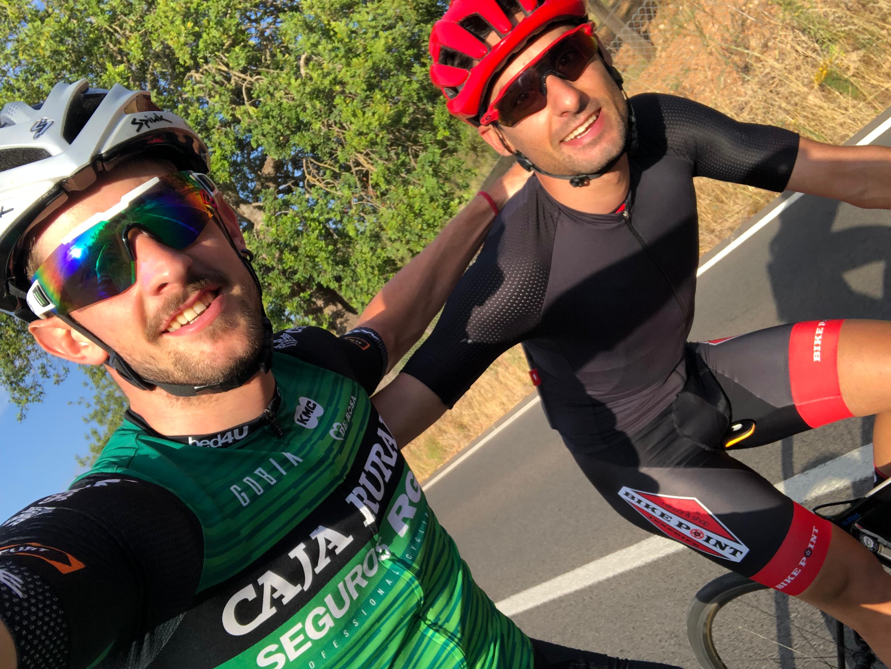James and Xavier taking a smiling cycling selfie on a sunny road, both in full kit during a guided ride in Mallorca